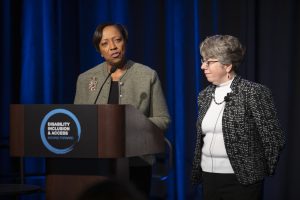 An African-American woman with short hair and a tan jacket speaks from a podium. Beside her, a white woman with short hair and glasses, wearing a black and white jacket looks on.