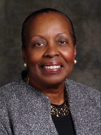 Claudette Lewis headshot description: A dark-skinned Black woman smiles at the camera. She is wearing a black top, a gray blazer, and pearl earrings. Her hair is pulled back. 