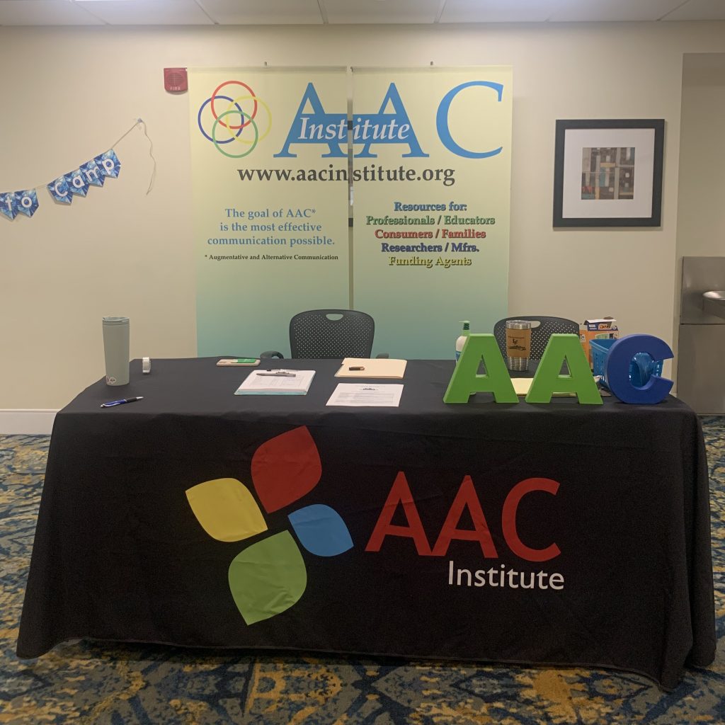 Display table of AAC Institute with AAC Institute logo. Behind the table is a standing banner that reads AAC Institute, www.aacinstitute.org. Lefthand side of banner reads The goal of AAC (augmentative and alternative communication) is the most effective communication possible. Righthand side of banner reads Resources for: Professionals/Educators, Consumers/Families, Researchers/Mfrs, Funding Agents.
