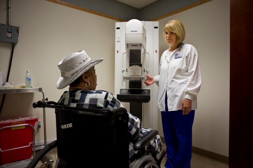 A female patient in a wheelchair talking to a female doctor. Behind the doctor is an accessible mammogram machine.