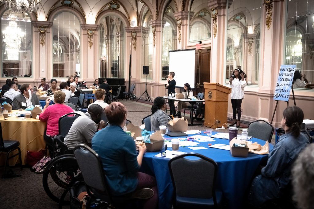 Image of participants sitting at round tables, listening to disability advocate Amanda Neatrour speak at the podium.