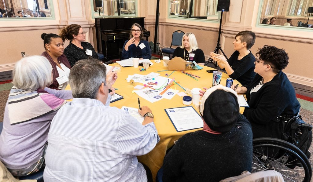 A group of nine disability advocates and allies sit around a round table and discuss potential solutions to address barriers to health-care access for people with disabilities.